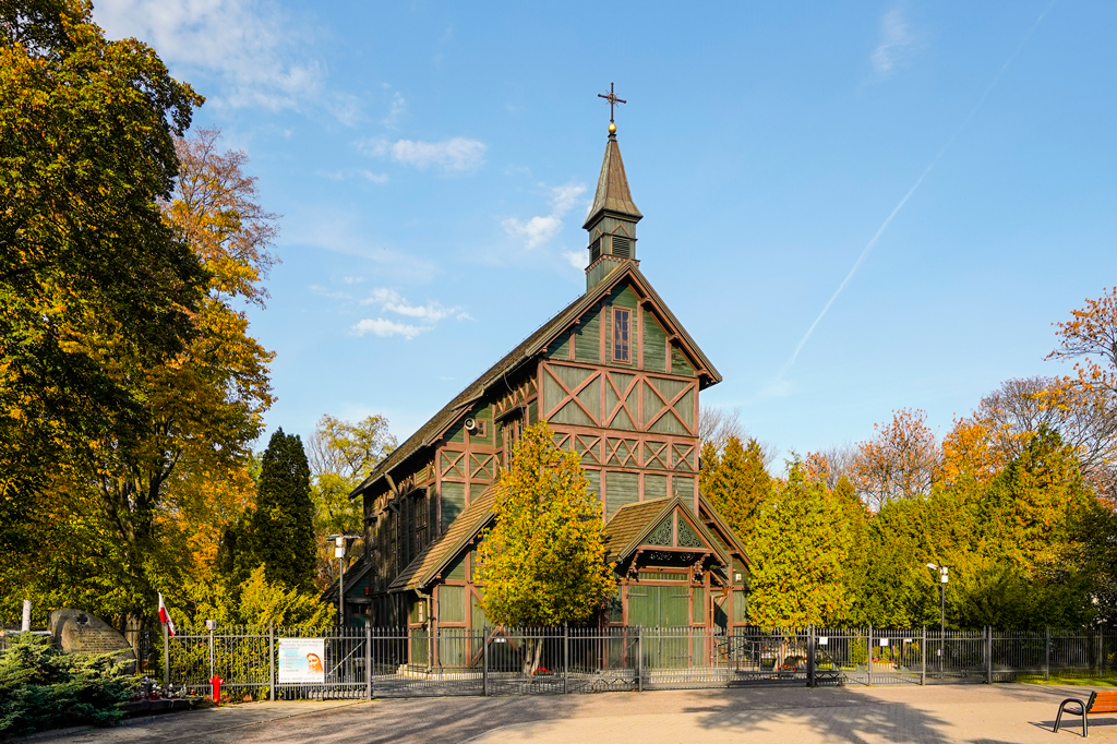 Drewniany kościół otoczony metalowym ogrodzeniem. Dookoła rosną drzewa. Słoneczny dzień, jesień.