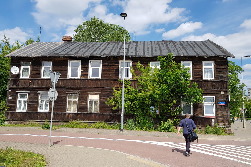 Drewniany dom przy ulicy Biruty 18, dookoła rosną drzewa, mężczyzna przechodzi przez ulicę, słoneczny dzień, lato.