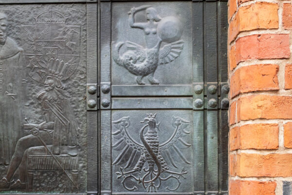 Fragment metalowych drzwi wejściowych do Archikatedry św. Jana, w drzwiach wykute różne postacie, między innymi syrenka i orzeł.