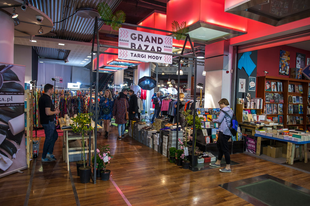 Wnętrze zabytkowego domu towarowego podczas targów mody „Grand Bazar”. Nad wejściem do strefy wystawienniczej wisi szyld z napisem „GRAND BAZAR TARGI MODY”. Wewnątrz widoczne są stoiska z ubraniami na wieszakach oraz regały z książkami po prawej stronie. Między stoiskami przechadzają się klienci. Wnętrze oświetlone jest nowoczesnymi, czerwonymi plafonami sufitowymi.