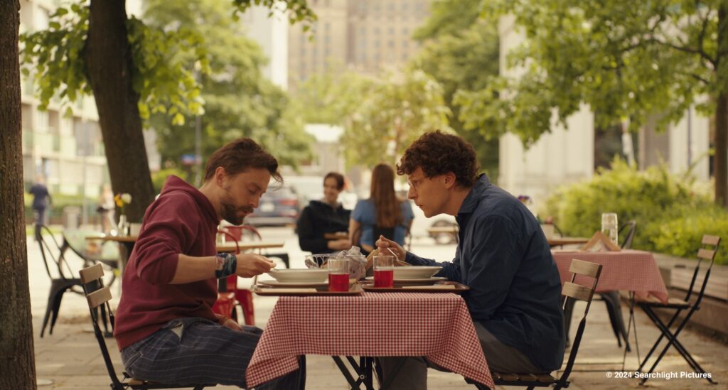 Dwóch mężczyzn siedzi przy stoliku, jedzą i rozmawiają, w tle inne stoliki, drzewa, dzień, lato.