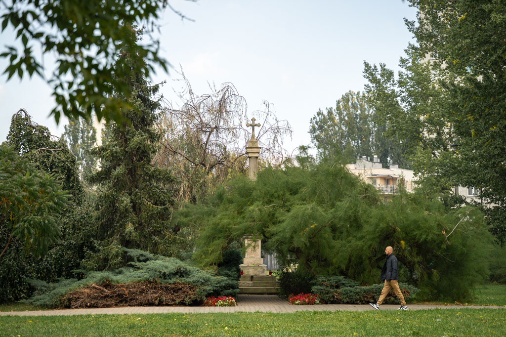 Kamienna kolumna, na jej szczycie krzyż, dookoła drzewa i krzewy, ścieżką spaceruje mężczyzna, dzień, lato.
