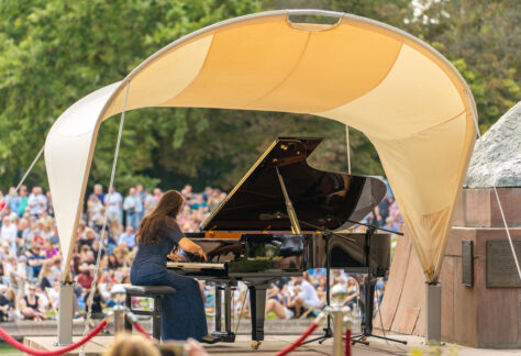 Koncert chopinowski w Łazienkach Królewskich, kobieta siedzi przy fortepianie, dookoła siedzą ludzie, w tle drzewa, dzień, lato.