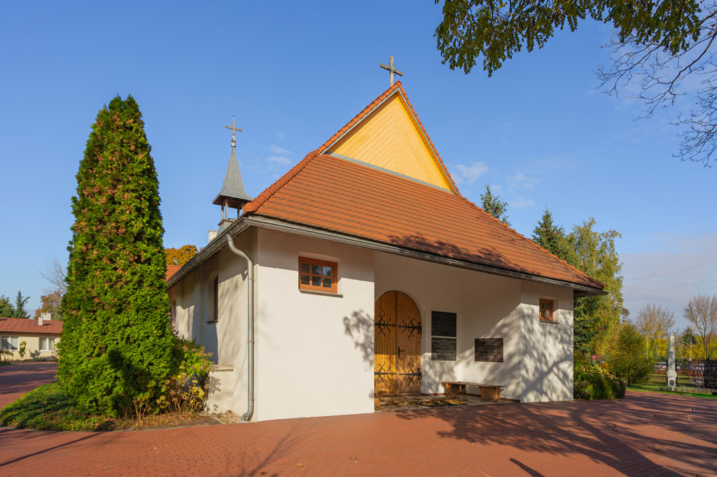 Mały, pomalowany na biało kościół z czerwonym dachem, dookoła rosną drzewa, słoneczny dzień, lato.