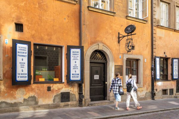 Widok na wejście do Muzeum Farmacji, nad wejściem metalowy szyld. Chodnikiem idą dwie kobiety.