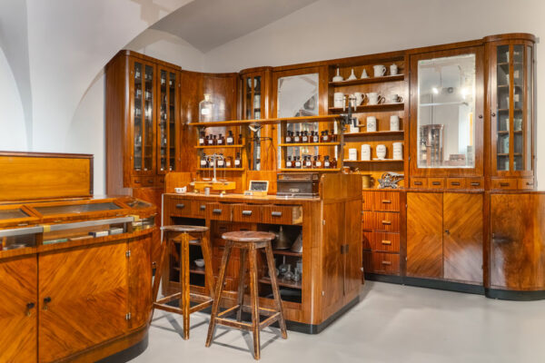 Sala ekspozycyjna w Muzeum Farmacji, drewniane regały i szafki. Na półkach dużo szklanych buteleczek i innych pojemników po lekach.