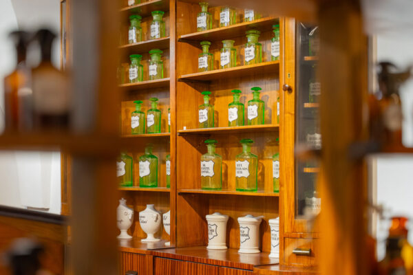 Muzeum Farmacji, na drewnianym regale stoją zielone przeźroczyste butelki z białymi etykietami oraz białe pojemniki na lekarstwa