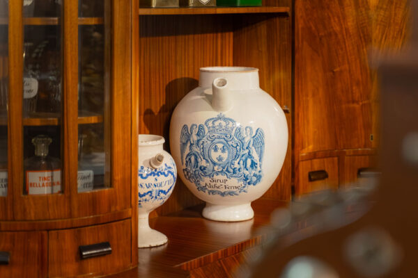 Muzeum Farmacji, na drewnianym regale stoją białe pojemniki na lekarstwa, za drzwiczkami szklane buteleczki.