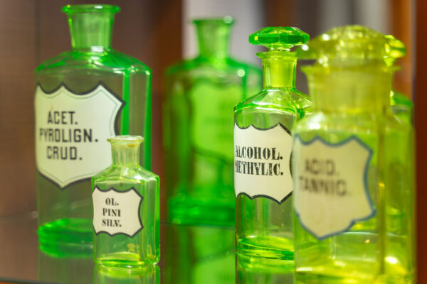 Eksponaty w Muzeum Farmacji, zielone przeźroczyste butelki, na białych etykietach czarne napisy.