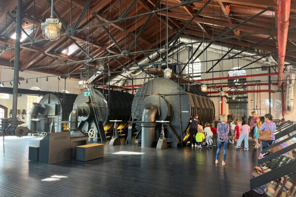 Sala ekspozycyjna Muzeum Gazowni Warszawskiej, na środku pomieszczenia stoją trzy duże czarne zbiorniki, obok grupa dzieci z opiekunami, po prawej stronie ekrany multimedialne.