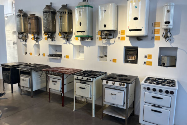 Sala ekspozycyjna Muzeum Gazowni Warszawskiej, na podłodze stoją dawne kuchenki, na ścianie wiszą dawne piecyki gazowe.