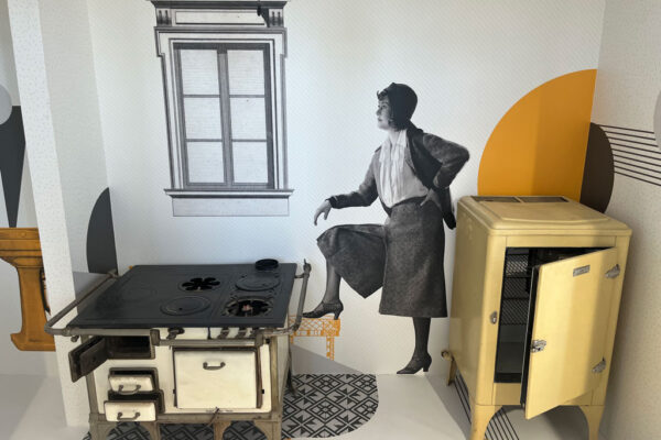 Sala ekspozycyjna Muzeum Gazowni Warszawskiej, pod ścianą dawna kuchenka oraz lodówka, na ścianie namalowana postać kobiety oraz okno.