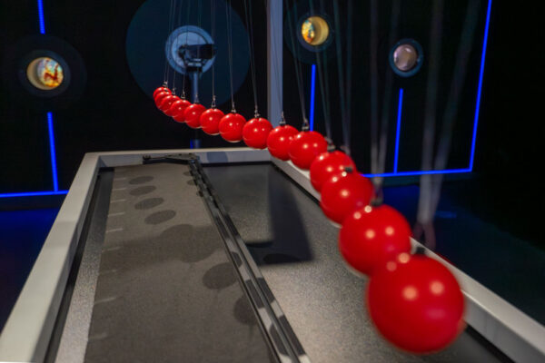 Bliskie ujęcie rzędu czerwonych kul zawieszonych na cienkich linkach, tworzących wahadło falowe. Kule są w ruchu, co powoduje ich rozmycie na pierwszym planie, układając się w kształt fali nad ciemnym blatem. W tle widać ciemną ścianę z podświetlonymi na niebiesko liniami i okrągłymi wizjerami.