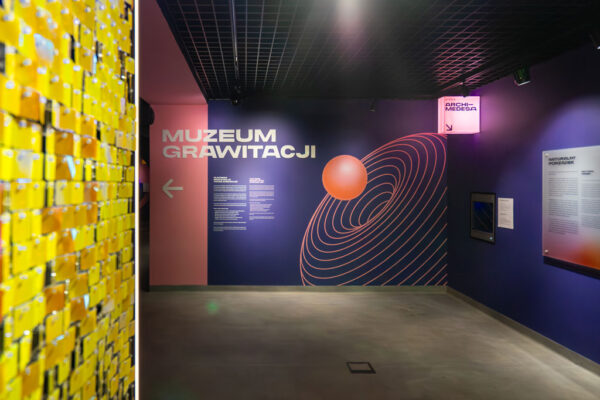 Nowoczesne wnętrze muzeum. Na fioletowej ścianie widnieje duży biały napis „MUZEUM GRAWITACJI” oraz grafika przedstawiająca pomarańczową kulę wpadającą w lej zakrzywionej czasoprzestrzeni. Po lewej stronie fragment ściany wyłożony błyszczącymi żółtymi płytkami.