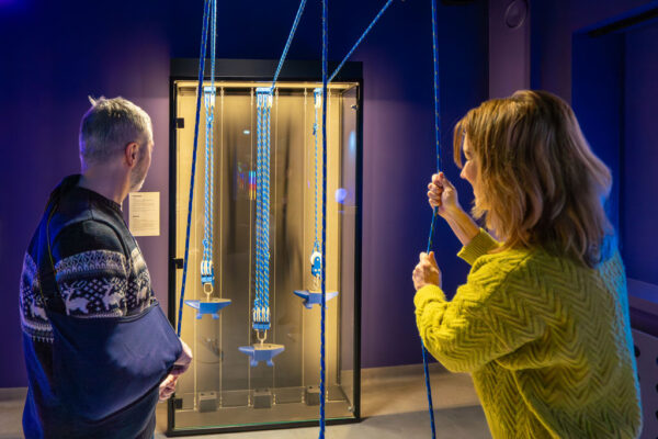 Kobieta w żółtym swetrze i mężczyzna w ciemnym swetrze z ortezą na ręku stoją przy przeszklonej gablocie. Wewnątrz gabloty znajdują się trzy kowadła zawieszone na niebieskich linach z różną liczbą krążków (wielokrążki). Kobieta trzyma za linę, testując działanie mechanizmu.