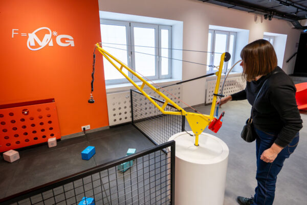 Kobieta operuje modelem żółtego żurawia (dźwigu) na białym postumencie. Na pomarańczowej ścianie w tle widnieje wzór fizyczny (siła ciężkości). Na podłodze leżą kolorowe klocki, które można podnosić za pomocą magnesu przymocowanego do ramienia żurawia.