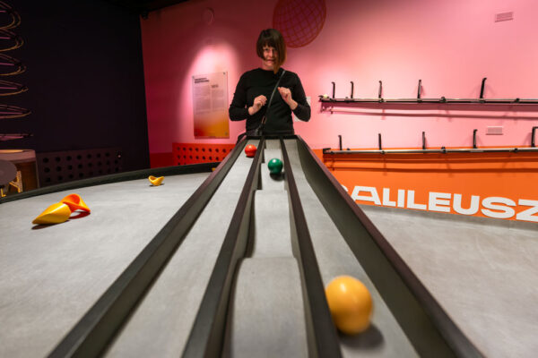 Kobieta w czarnym ubraniu stoi przy długim, szarym torze zjazdowym z kilkoma rynnami. W rynnach znajdują się kolorowe kule w różnym stadium ruchu. Na pomarańczowej ścianie w tle widnieje fragment napisu „GALILEUSZ”.
