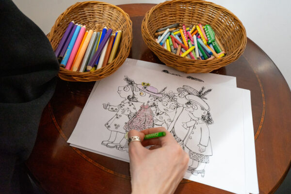 Zbliżenie na dłoń osoby kolorującej zieloną kredką rysunek satyryczny przedstawiający grupę postaci. Na drewnianym blacie obok kartki stoją dwa wiklinowe koszyczki wypełnione kolorowymi kredkami i pastelami.