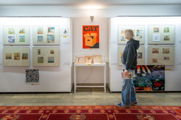 Wnętrze galerii. Na białych ścianach wiszą ramy z wieloma drobnymi rysunkami satyrycznymi. W centralnym punkcie, nad stolikiem z eksponatami, widnieje duży pomarańczowy plakat z napisem „CAT”, przedstawiający kota w kartonowym pudełku. Po prawej stronie bokiem stoi osoba w ciemnej bluzie i jeansach, oglądająca ekspozycję. Na podłodze leży wzorzysty, bordowy dywan.