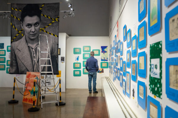 Mężczyzna stoi tyłem w sali wystawowej, przyglądając się ścianie zapełnionej licznymi małymi rysunkami w niebieskich oprawach. Po lewej stronie wisi wielkoformatowy, czarno-biały portret kobiety, przysłonięty częściowo przez srebrną drabinę malarską zabezpieczoną żółto-czarnymi łańcuchami.