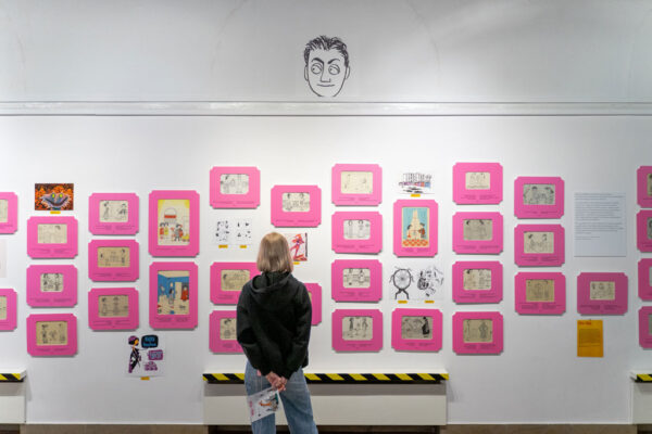 Kobieta o blond włosach ogląda ścianę z ekspozycją prac w jaskraworóżowych, dekoracyjnych ramkach. Rysunki przedstawiają sceny z życia codziennego w stylu retro. Nad ramkami, bezpośrednio na białej ścianie, namalowana jest czarną kreską twarz mężczyzny o zdziwionym wyrazie.