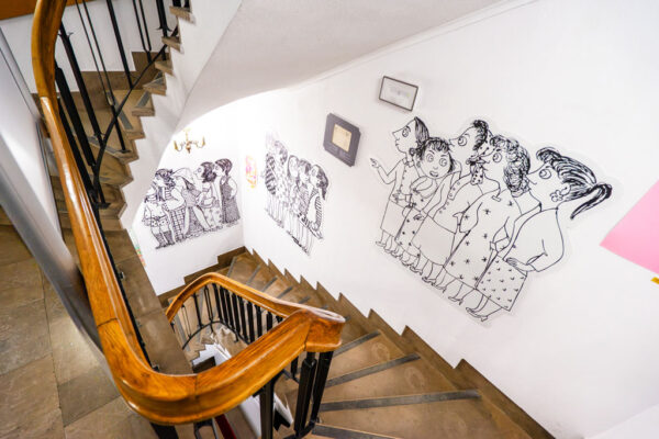 Widok z góry na kręte schody z drewnianą poręczą i metalową balustradą. Białe ściany wzdłuż schodów ozdobione są dużymi, czarno-białymi postaciami rysunkowymi przedstawiającymi grupy ludzi.