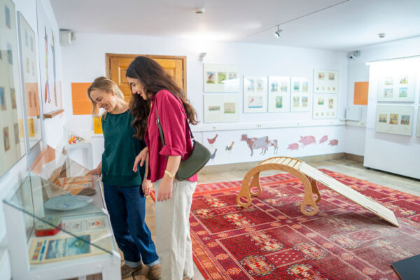 Wnętrze jasnej sali muzealnej, w której dwie kobiety pochylają się nad niską szklaną gablotą z eksponatami. Na białych ścianach wiszą kolorowe grafiki i ilustracje, a na dole ściany namalowane są sylwetki zwierząt gospodarskich. Na środku pokoju leży duży, czerwony dywan o orientalnym wzorze, na którym stoi drewniana zabawka w kształcie łuku z pochylnią.