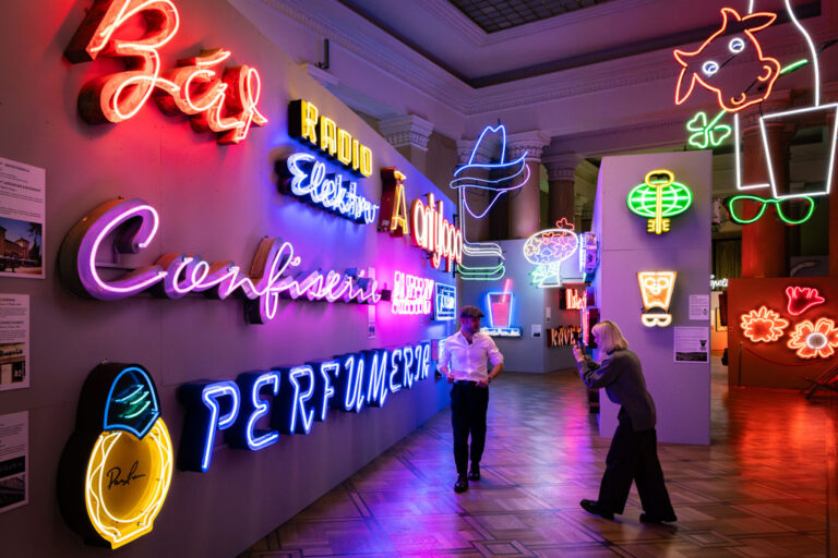 Muzeum Neonów, kobieta robi mężczyźnie zdjęcie na tle kolorowych neonów zawieszonych na ścianach.