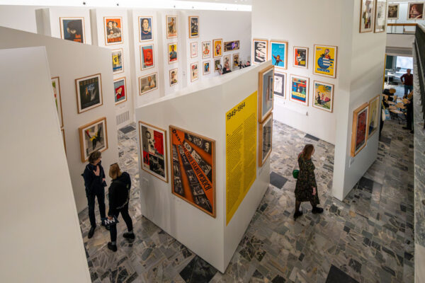 Widok z góry na wielopoziomową przestrzeń wystawienniczą. Białe ścianki działowe tworzą labirynt, na którym gęsto eksponowane są kolorowe polskie plakaty. W centralnym punkcie widoczna żółta sekcja z datami 1949–1964. Pomiędzy ściankami spacerują zwiedzający.