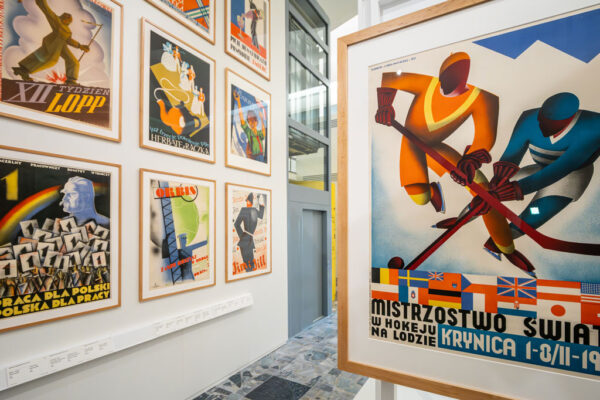 Fragment wystawy prezentujący plakaty w drewnianych ramkach. Na pierwszym planie po prawej dynamiczny plakat hokejowy z napisem „Mistrzostwa Świata w hokeju na lodzie, Krynica 1-8/II-1931”. W tle inne grafiki, w tym plakat z logotypem biura podróży Orbis.