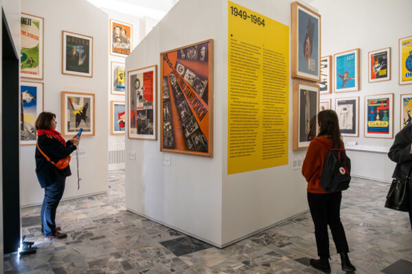 Dwie osoby zwiedzające sekcję poświęconą polskiemu plakatowi z lat 1949–1964. Na białej ścianie działowej umieszczono duży żółty panel z tekstem kuratorskim w języku polskim i angielskim. Wokół wiszą plakaty z okresu socrealizmu oraz wczesnej Polskiej Szkoły Plakatu.