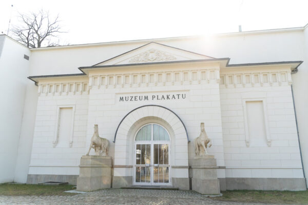 Klasycystyczna, biała fasada Muzeum Plakatu w Wilanowie. Nad półkolistym, przeszklonym wejściem widnieje napis „MUZEUM PLAKATU”. Po obu stronach drzwi znajdują się rzeźby białych koni na podestach.