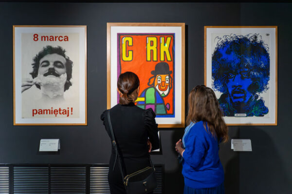 Trzy plakaty wiszące obok siebie na ciemnej ścianie. Od lewej: plakat „8 marca pamiętaj!” z czarno-białym zdjęciem mężczyzny z pianką do golenia, kolorowy plakat cyrkowy z klaunem i napisem „CYRK” oraz niebiesko-czarny portret Jimiego Hendrixa. Przed nimi stoją dwie tyłem odwrócone kobiety.