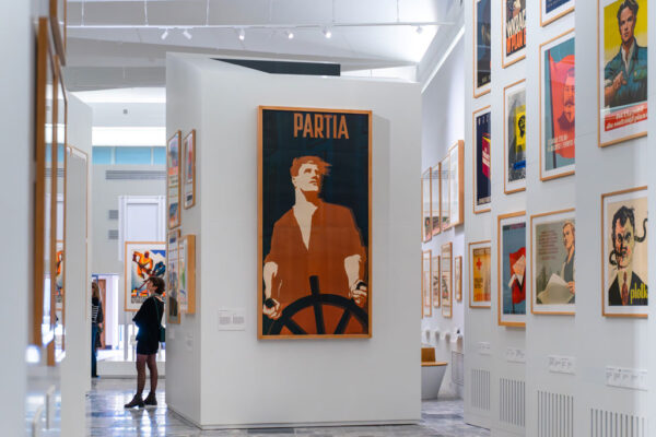 Centralny punkt kadru zajmuje duży plakat na białej ścianie przedstawiający marynarza przy sterze na ciemnym tle z napisem „PARTIA”. Po lewej stronie kobieta w czarnej sukience przygląda się ekspozycji w głębi sali. W tle rzędy innych mniejszych plakatów.