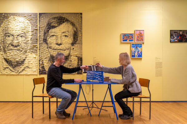 Kobieta i mężczyzna siedzą naprzeciwko siebie przy niebieskim stoliku na tle żółtej ściany. Uczestniczą w grze lub aktywności manualnej, wyciągając ręce w stronę niebieskiego obiektu na blacie. W tle widoczne są dwa wielkoformatowe portrety wykonane z karteczek (kobiety i mężczyzny) oraz trzy mniejsze, kolorowe obrazy w ramkach.