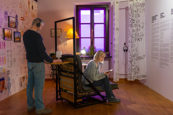 Scena na wystawie imitująca domowe wnętrze. Kobieta siedzi na rozłożonym fotelu-łóżku i czyta książkę. Obok niej stoi mężczyzna w słuchawkach, oparty o blat roboczy zamocowany do stelaża łóżka. W tle okno z fioletową szybą, zapalona lampka nocna oraz biała ściana gęsto pokryta czarnymi rysunkami i komiksami.