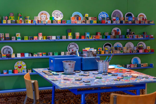 Zbliżenie na niebieski blat stołu warsztatowego, gęsto pokryty plamami z kolorowych farb. Na stole stoją metalowe kubki z pędzlami oraz niebieskie pojemniki z farbami w tubkach. W tle widać zieloną ścianę z rzędami pomalowanych przez zwiedzających klocków i talerzyków, tworzących barwną mozaikę.