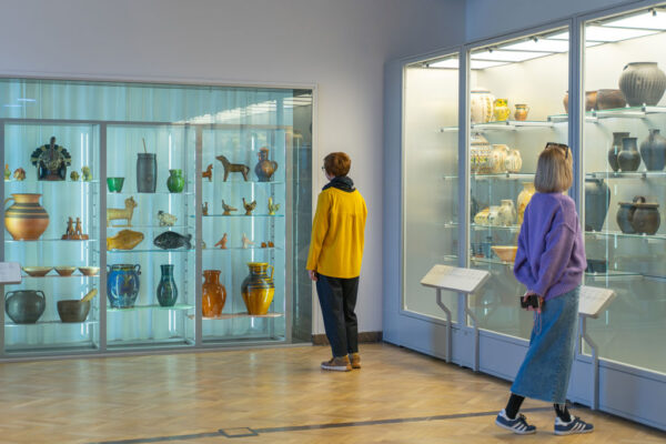 Dwie kobiety oglądają wystawę ceramiki w muzeum. Jedna, w żółtej bluzie, stoi przed dużą, wbudowaną w ścianę gablotą z naczyniami i figurkami. Druga, w fioletowym swetrze, podchodzi do kolejnej przeszklonej gabloty po prawej stronie, na której półkach stoją gliniane dzbany i wazy.