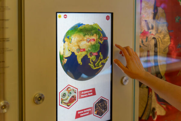 Zbliżenie na pionowy ekran dotykowy wbudowany w jasną ścianę gabloty w muzeum. Na ekranie wyświetla się interaktywny model kuli ziemskiej z zaznaczonym kontynentem azjatyckim i czerwonymi znacznikami, takimi jak „Indie” czy „Japonia”. Pod globusem widoczne są kafelki z napisami „Azjatycki Nowy Rok” oraz „Inne święta świateł”. Z prawej strony widoczna jest dłoń uniesiona w kierunku ekranu. W tle, za szybą gabloty, majaczy fragment tradycyjnego, azjatyckiego wizerunku.