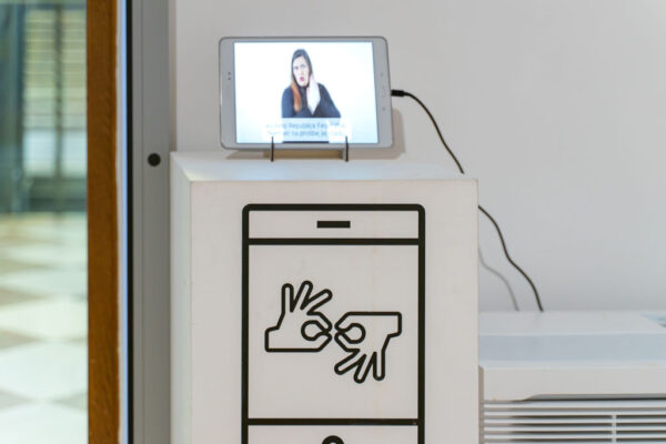 Fragment muzealnej ekspozycji dostosowanej dla osób głuchych. Na białym postumencie stoi tablet, na którego ekranie wyświetlane jest nagranie z tłumaczką Polskiego Języka Migowego (PJM) oraz napisy w języku polskim. Na froncie postumentu znajduje się duża, czarna ikona przedstawiająca smartfon, na którym widnieje międzynarodowy symbol migania.