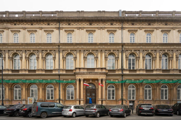 Szerokie ujęcie trzykondygnacyjnego budynku Państwowego Muzeum Etnograficznego. Nad centralnie położonymi, drewnianymi drzwiami wejściowymi widnieje napis „PAŃSTWOWE MUZEUM ETNOGRAFICZNE”, a obok wejścia wiszą dwie flagi: Polski oraz Mazowsza. Przed budynkiem, wzdłuż krawężnika, zaparkowany jest rząd samochodów osobowych, a nad parterem rozciągnięta jest zielona siatka ochronna.