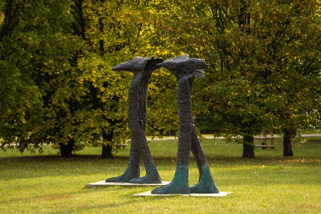 Dwie rzeźby postaci o ludzkich ciałach i zwierzęcych głowach ustawione na trawie, dookoła rosną drzewa, dzień, jesień.