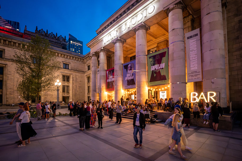 Plac przed wejściem do Baru Studio, ludzie spacerują, inni siedzą przy stolikach, wieczór, lato.