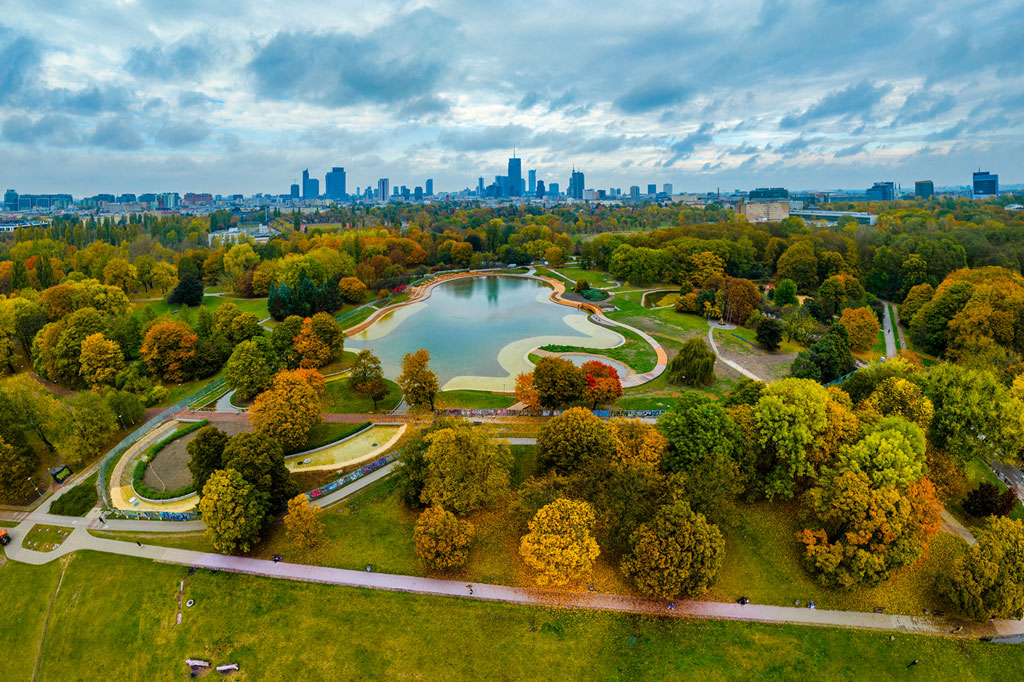 Widok z góry na Pole Mokotowskie, na pierwszym planie staw otoczony drzewami, w tle panorama centrum Warszawy, pochmurny dzień, jesień.