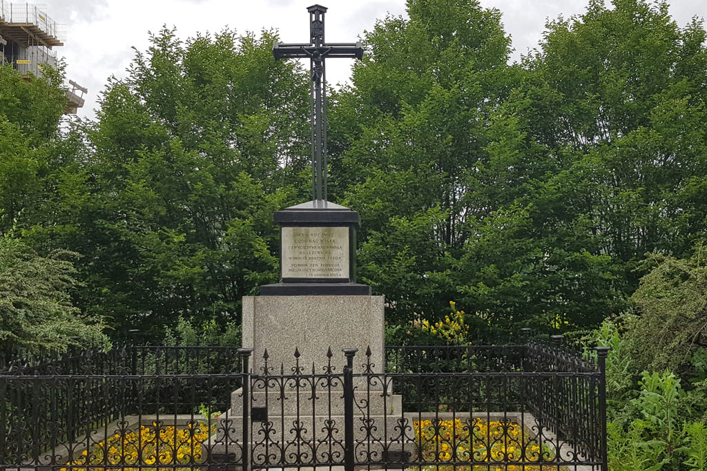 Za niskim metalowym ogrodzeniem na murowanym postumencie stoi metalowy krzyż. Dookoła rosną drzewa i krzewy.