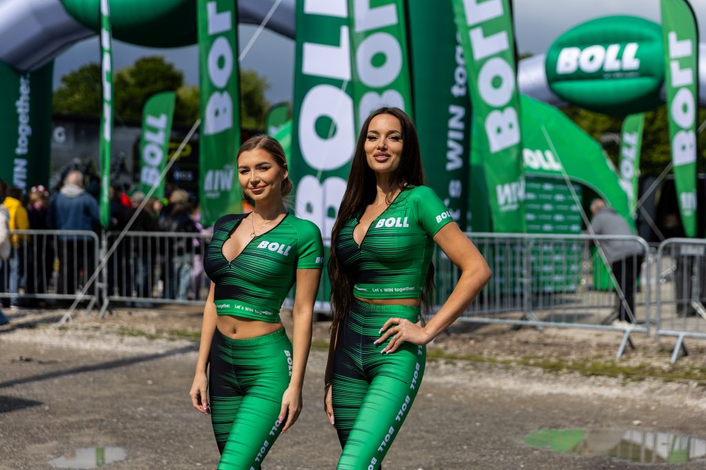 Two smiling women in matching green and black sportswear with the BOLL brand logo visible. In the background, you can see numerous green flags and advertising banners of the BOLL brand, an inflatable gate and a promotional balloon.