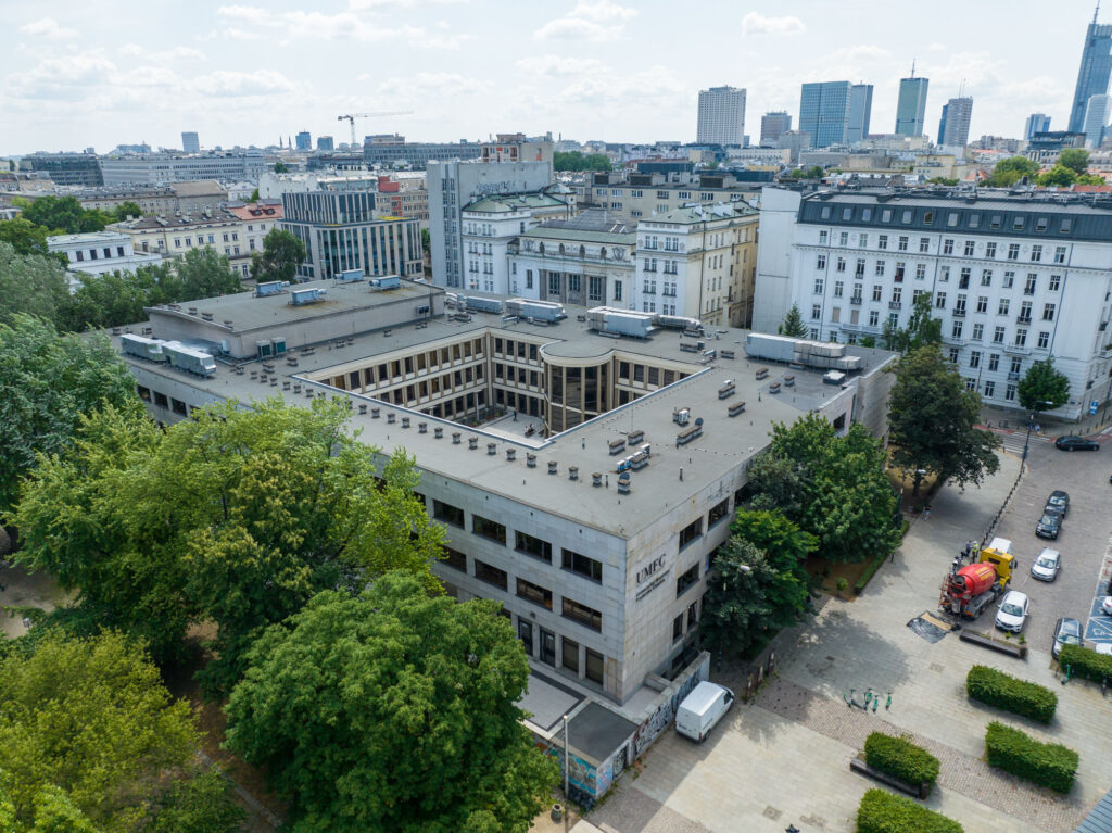 Widok z góry na zabudowę centrum Warszawy, z modernistycznym kompleksem budynków na pierwszym planie i panoramą wieżowców w tle.