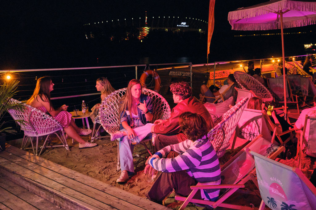 Grupa ludzi bawi się w klubie na bulwarach wiślanych, w tle Wisła, stadion PGE Narodowy, noc, lato.