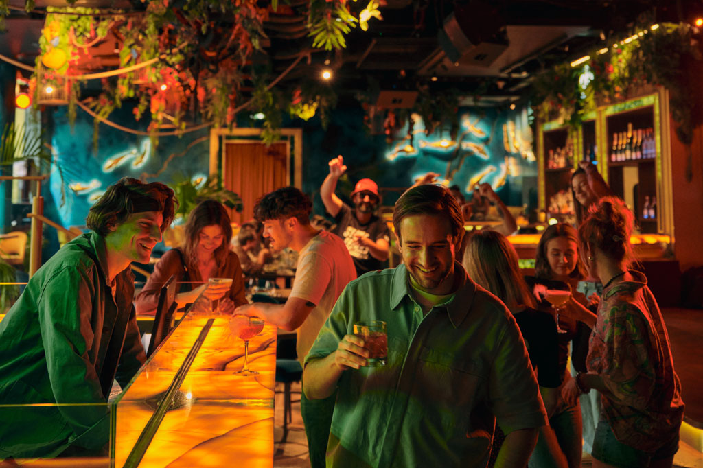 Wieczór w barze, ludzie siedzą przy stolikach, piją, rozmawiają.