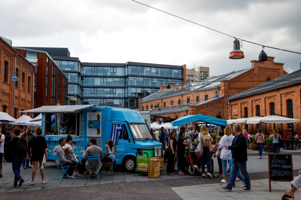 Zlot food trucków w Centrum Praskim Koneser, ludzie spacerują, inni siedzą przy stolikach, pochmurny dzień, lato.
